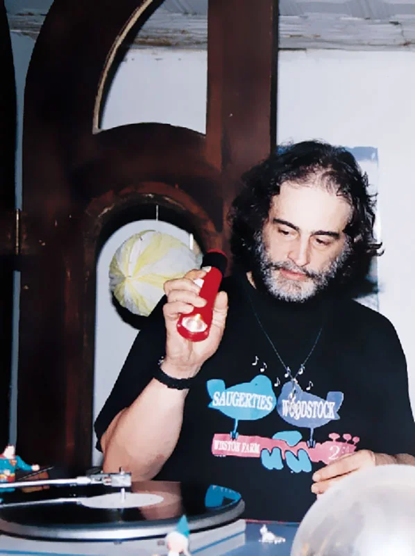 David Mancuso at the turntables on Avenue A, grey-bearded, in a black T-shirt, looking down at a record.