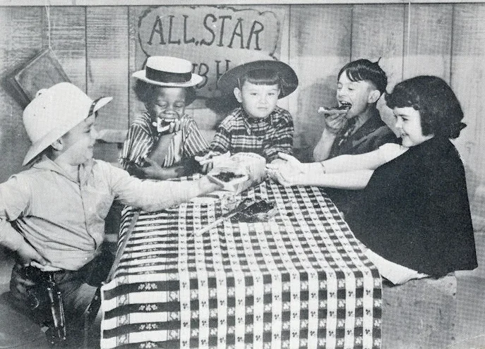 Black-and-white film still: five children at a table with a checked cloth under a hand-lettered ALL STAR sign, sharing food; one wears a straw boater, one a cowboy hat.