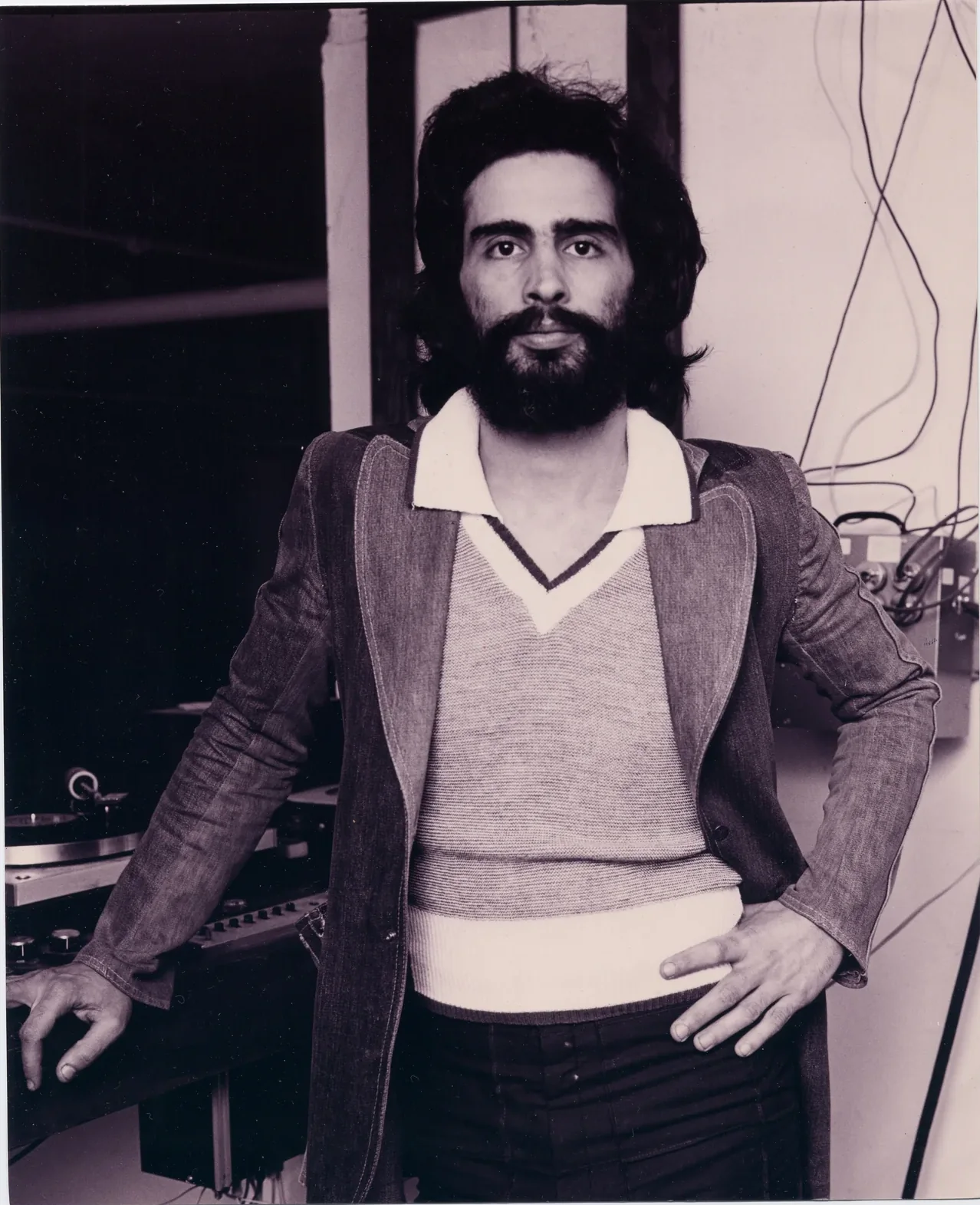 Black-and-white portrait of David Mancuso in the Prince Street loft: long dark hair and beard, a jacket over a V-neck, hands on his hips.