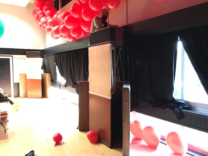 Two tall wooden Klipschorn loudspeakers set up in a bright hall, red balloons crowding the ceiling above them.