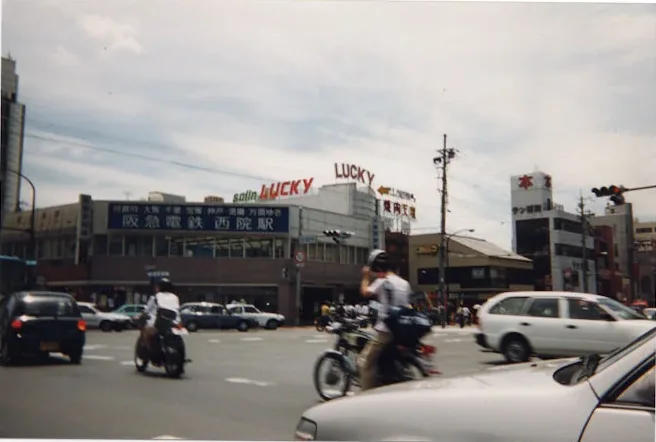 Colour snapshot of a street corner in Japan, a sign reading LUCKY above the shops, motorbikes in the road.
